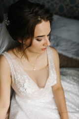 Portrait of a bride sitting indoors in her wedding dress. She looks at the camera with a calm, confident expression. Soft light highlights the lace details, veil, and floral bracelet, creating 