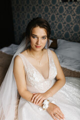 Portrait of a bride sitting indoors in her wedding dress. She looks at the camera with a calm, confident expression. Soft light highlights the lace details, veil, and floral bracelet, creating 