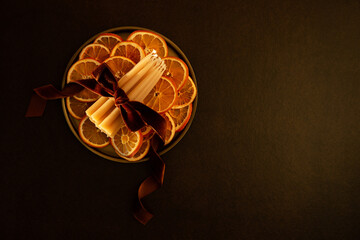 Décoration de Noël, tranches d’oranges séchées, fagot de bougies en cire naturelle entouré d’un ruban en velours brun orangé avec un noeud, posé sur un plateau doré, fond brun foncé, vue du dessus