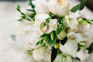 Close-up of wedding rings placed on a bouquet of white flowers. Soft natural light highlights the delicate petals, greenery, and golden bands, creating a romantic and elegant wedding detail shot.