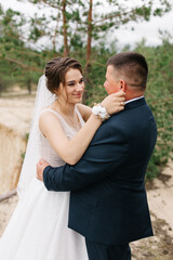 Fototapeta premium The bride gently embraces the groom, smiling and looking into his eyes. The forest background highlights sincere emotions, warmth, and intimacy of this special wedding moment.