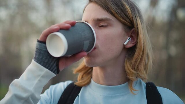 teen enjoying beverage outdoors calmly, casual youth with earbuds holding coffee in peaceful park setting, teenager calmly sipping coffee while listening through earbuds in outdoor park scene