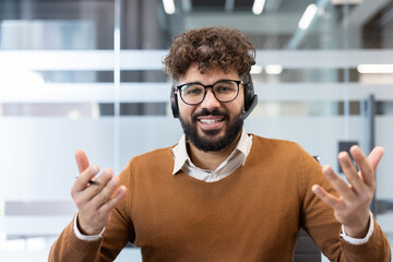 Obraz premium Smiling bearded man working as customer service in an office, wearing a headset and glasses, actively communicating and providing support during a video call