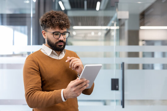 Young businessman with curly hair and glasses stands in a bright modern office, focused on a digital tablet while reviewing data, messaging, and planning strategy - Powered by Adobe