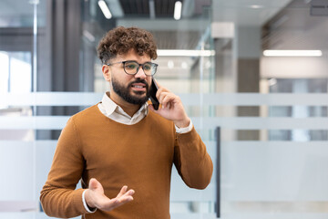 Young businessman standing in a contemporary office setting, having an important conversation on his mobile phone while actively gesturing with one hand, communicating business ideas