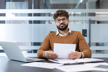 Young bearded man with eyeglasses sitting at a desk in a modern office, holding a blank document, laptop and papers on the table, looking directly at the camera with a serious expression