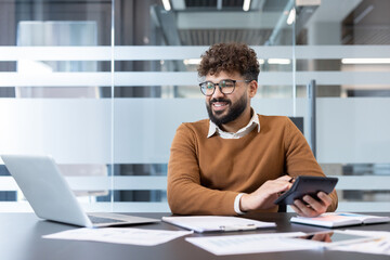 Young man calculating finances and paperwork at his desk, smiling and looking satisfied while working on business projects in a contemporary office environment