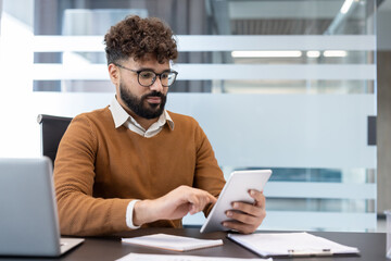 Young businessman with curly hair and glasses working on a digital tablet at his desk, analyzing important financial data and information in a modern business office environment