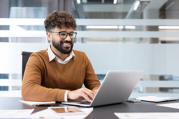 Young smiling businessman with glasses and beard working efficiently on his laptop at a modern corporate office desk, typing and managing his digital tasks with positive engagement and focus