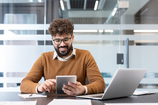 Young professional man with curly hair and glasses working remotely, interacting with a digital tablet and laptop, collaborating in a modern office environment