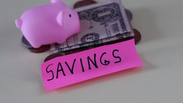 Un primer plano de un cerdo de color rosado con una nota con la palabra ahorros y rodeado de dinero en un fondo de color amarillo claro