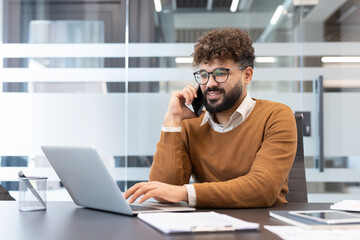 Young businessman multitasking, making a phone call while typing on a laptop, smiling at his desk in a modern corporate office environment with glass walls