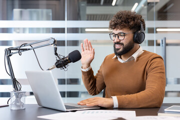 Young man wearing headphones and glasses, recording a podcast, speaking into a microphone and using a laptop, actively engaging with listeners or guests