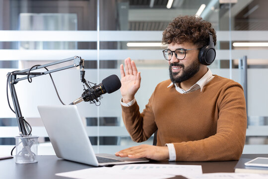 Young man wearing headphones and glasses, recording a podcast, speaking into a microphone and using a laptop, actively engaging with listeners or guests
