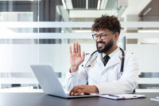 Young male doctor in a white coat and stethoscope smiling and waving during a virtual medical appointment or healthcare video call, on a laptop in a clinic office - Powered by Adobe