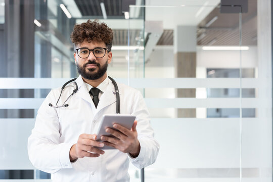 Indian doctor in lab coat and stethoscope holding digital tablet, looking at camera while standing in a modern clinic or hospital, representing digital healthcare and medical technology