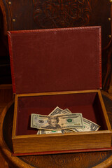 US dollar bills inside an open wooden box with a lid and red lining. The object is on a dark wooden stool. Concept of finance, saving, wealth, secrecy, profit and illicit business.