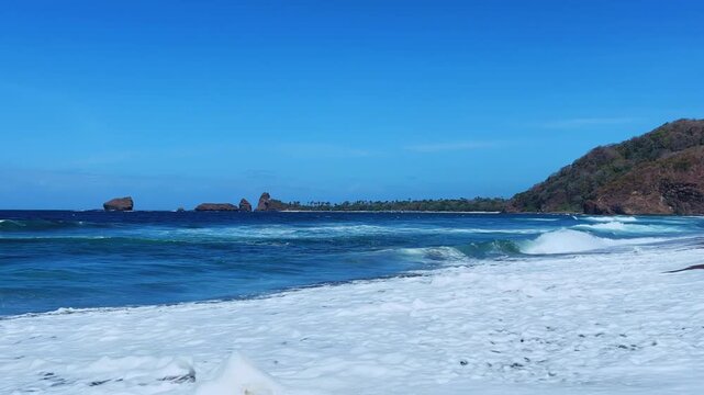The sea in Jember, East Java. Indonesia. Watu Ulo beach with dark sand, mountains and coral rocks towering above the beach. The raging blue Indian ocean on a sunny day. 4К