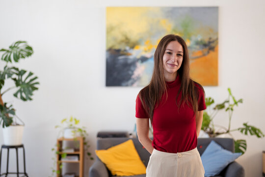 A young woman stands in a modern living room, smiling at the camera. She wears a red shirt and light-colored pants, with plants and artwork in the background.