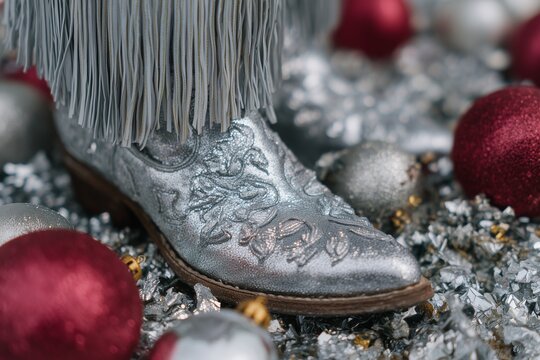 Silver cowboy boots adorned with intricate designs and fringes, surrounded by festive ornaments, creating a vibrant holiday atmosphere with a touch of western flair