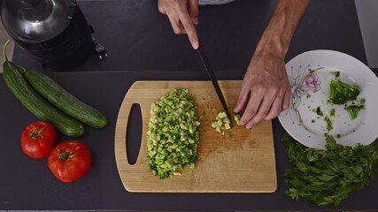 A detailed overhead shot captures a man's hands meticulously dicing fresh broccoli on a wooden board, surrounded by healthy ingredients like tomatoes and cucumbers. - Powered by Adobe