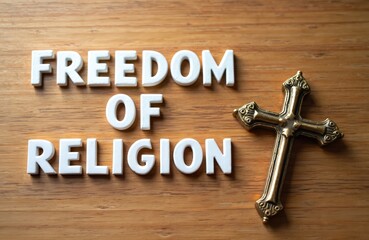 White letters spell Freedom of Religion on wood background. Bronze Christian cross rests beside words. Image symbolizes spiritual liberty, human rights, and faith belief protection.