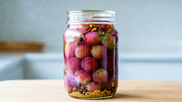 Glass jar filled with pickled grapes in vinegar brine, standing on a wooden table in bright kitchen, preserved fruit for garnish, appetizer, cheese board, fermentation, culinary trend, pantry storage
