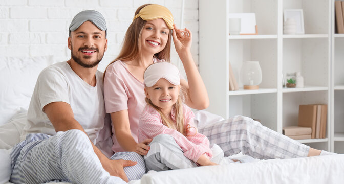 Happy parents and their little daughter with sleeping masks sitting in bedroom