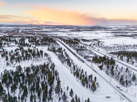 Aerial view of a train slicing through the snowy landscape, with the soft glow of the horizon kissing the distant mountains, Kiruna, Norrbotten County, Sweden.