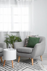 Cozy grey armchair with cushions and jug of water on table in interior of bright living room