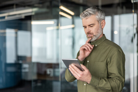 Experienced man holding contemporary digital tablet, thinking about strategy or problem-solving, dressed in a olive shirt in a modern office, representing business innovation and technology - Powered by Adobe