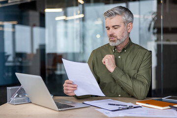 Mature executive focused on paperwork and data analysis, sitting at a modern office desk with a laptop and various documents, embodying diligence and corporate responsibility