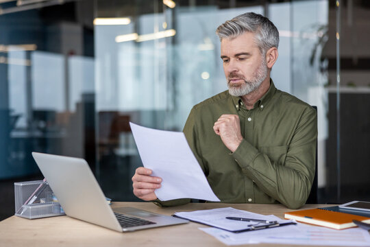 Mature executive focused on paperwork and data analysis, sitting at a modern office desk with a laptop and various documents, embodying diligence and corporate responsibility