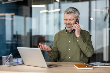 Mature man working in modern office, smiling professional sitting at desk with laptop, having phone conversation and gesturing, communicating good news or negotiating a deal