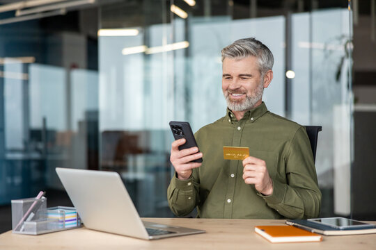 Mature man with gray hair smiling as he completes a secure online payment on his smartphone using a credit card, seated at a desk with laptop in a modern office setting