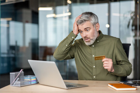 Shocked mature man looking at his laptop screen while holding a credit card, reacting to an online fraud, debt, or financial problem at his office desk