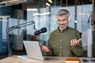 Mature man wearing headphones and using a studio microphone with a laptop, broadcasting or recording an online podcast from a professional office environment, actively gesturing while speaking