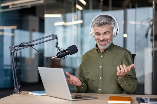 Mature man wearing headphones and using a studio microphone with a laptop, broadcasting or recording an online podcast from a professional office environment, actively gesturing while speaking