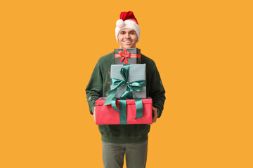Handsome young man in Santa hat holding Christmas gifts on yellow background