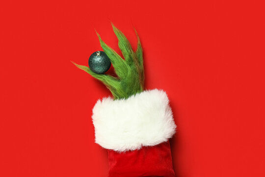 Hairy hand of green creature in Santa costume with Christmas ball on red background