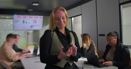 Smiling businesswoman standing in office meeting room holding smartphone, diverse colleagues working on laptops with data charts on screen in background