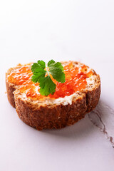 Fresh homemade canape made from full grain baguette, butter and salmon caviar deocrated with aromatic cilantro leves on white marble table macro view.
