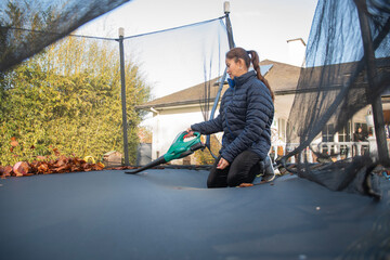 Woman using a leaf blower to clear dry autumn leaves from a backyard trampoline, performing seasonal yard work to maintain outdoor play equipment and tidy the garden area