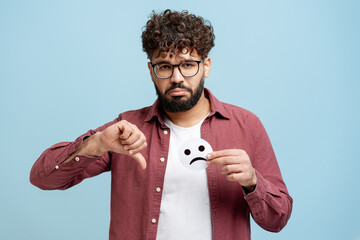Disappointed man holding a sad emoticon and gesturing thumbs down, indicating disapproval