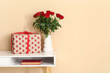 White desk with bouquet of roses, gift box and book near beige wall. Valentine's Day celebration
