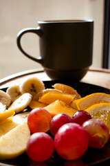 Close-up breakfast still life with sliced fresh fruits on a plate and a blurred “Coffee House” mug in the background. Bright side light creates glowing highlights and a cozy, inviting mood.