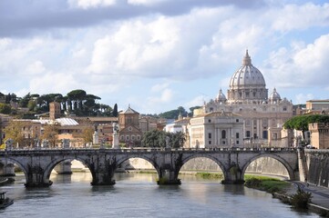 View of St. Peter’s Basilica and Ponte Sant’Angelo in Rome, with the Tiber River reflecting the historic bridge and the iconic dome under soft daylight and dramatic clouds.