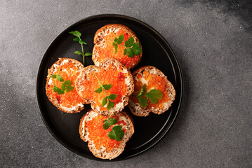 Fresh homemade canape made from full grain baguette, butter and salmon caviar deocrated with aromatic cilantro leaves on black ceramic plate top view.