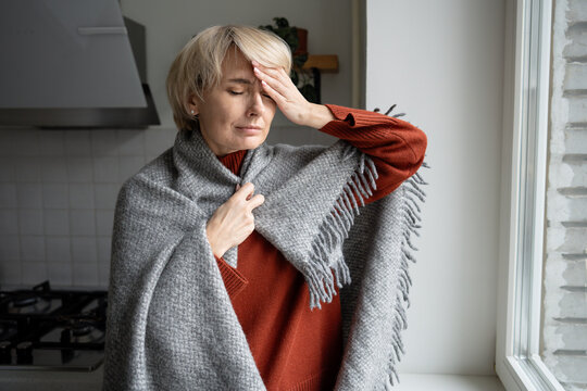Middle aged woman in wool plaid touching forehead with painful expression, suffering from heavy migraine and high temperature, signs of cold and flu, health problems during cold season and infection.