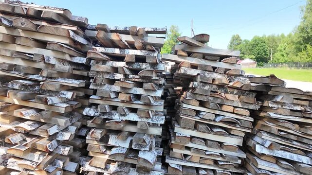 stacked birch firewood piles outdoors, sunlit summer sky, grassy yard with blue fence, closeup of peeling bark and split logs, rustic texture and seasonal